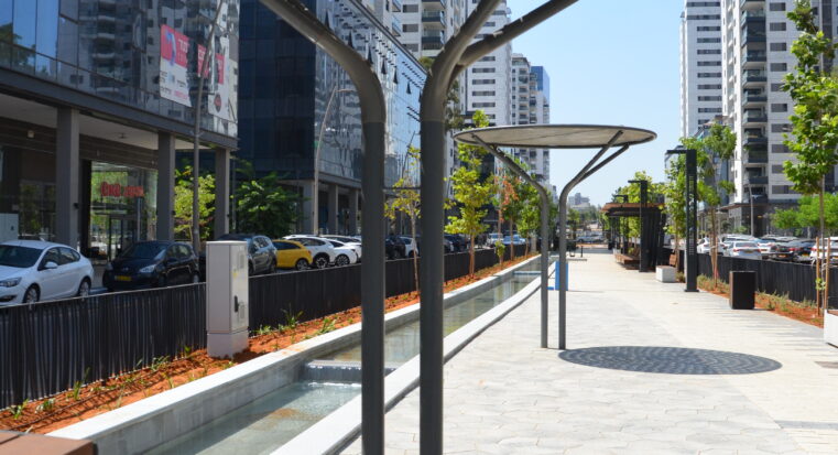 פרגולת יערה ממתכת עם גג עגול ומחורר, מותקנת בטיילת עירונית לצד תעלת מים