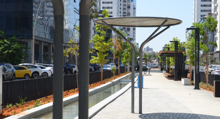 פרגולת יערה ממתכת עם גג עגול ומחורר, מותקנת בטיילת עירונית לצד תעלת מים