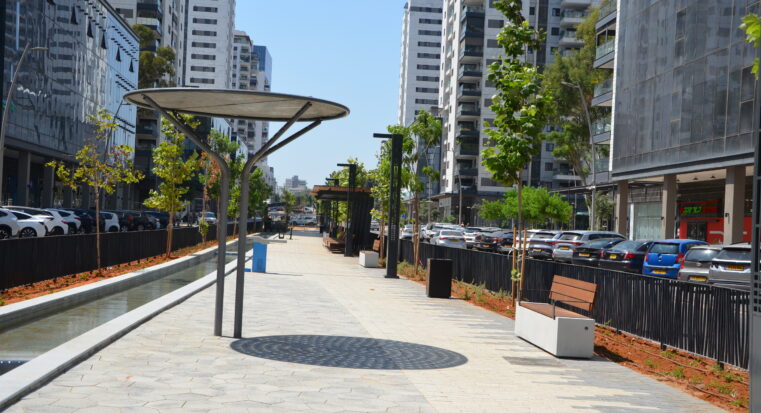 פרגולת יערה ממתכת עם גג עגול ומחורר, מותקנת בטיילת עירונית לצד תעלת מים