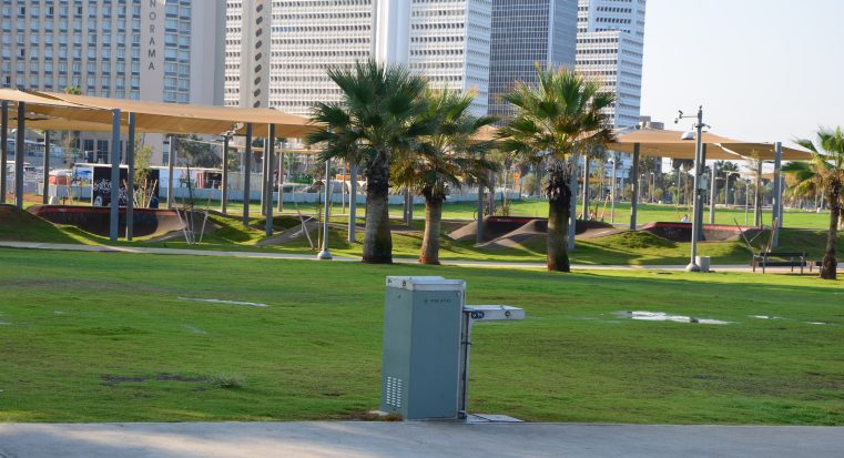 ברזיית דן של שחם אריכא, מותקנת במדשאה עירונית עם רקע של עצים ומגדלים בטיילת תל אביב